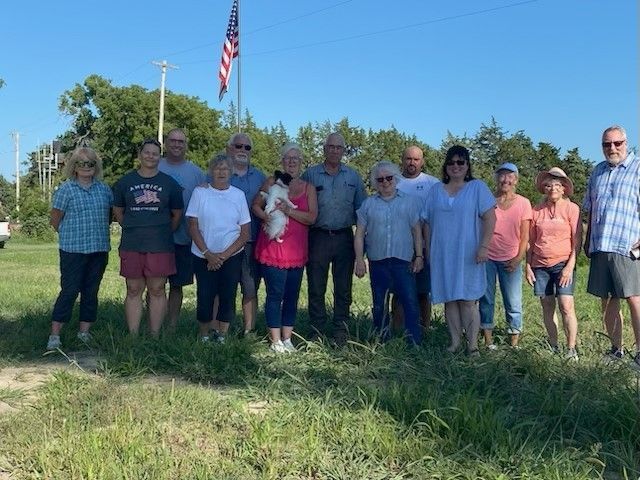 Group of people standing in a grassy field with an American flag in the background on a sunny day.