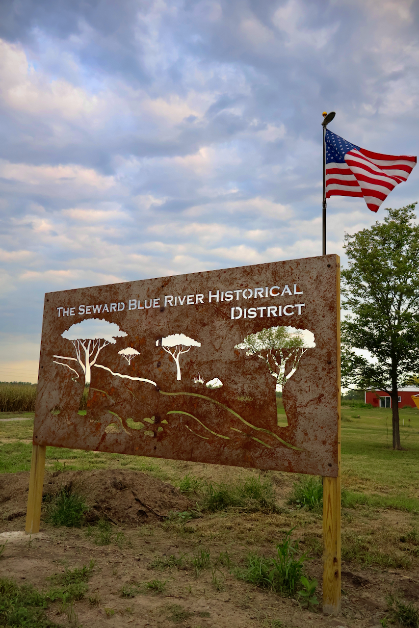 Sign for Seward Blue River Historical District with American flag and cloudy sky.