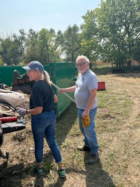Woman and man cleaning up an outdoor area; woman holding tool, man leaning on a bin.