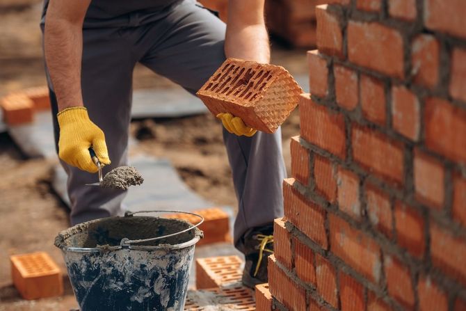 Worker laying red bricks and applying mortar to a wall with a trowel.