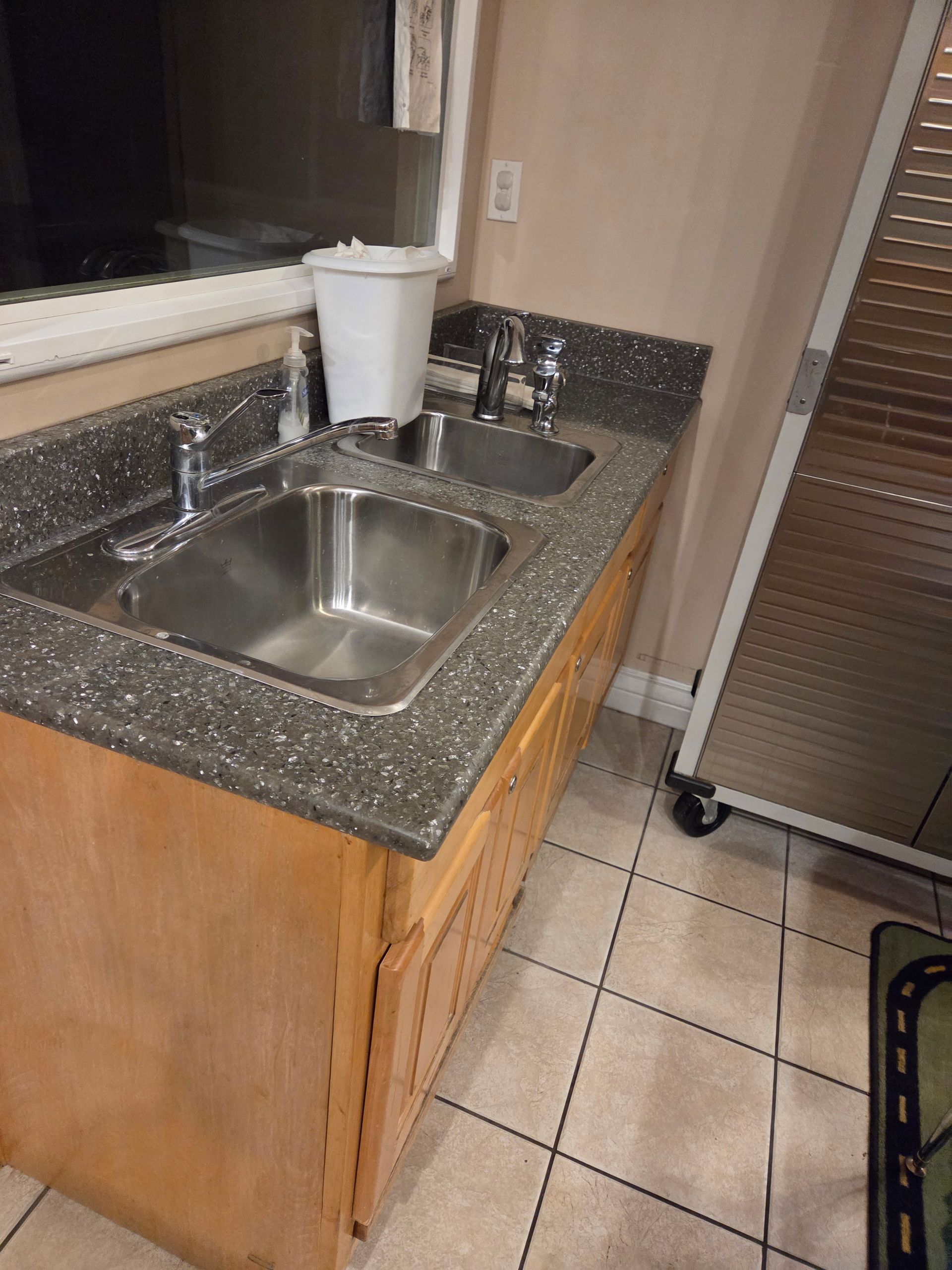 Kitchen sink with speckled countertop, wooden cabinet, tiled floor, and stainless steel cart nearby