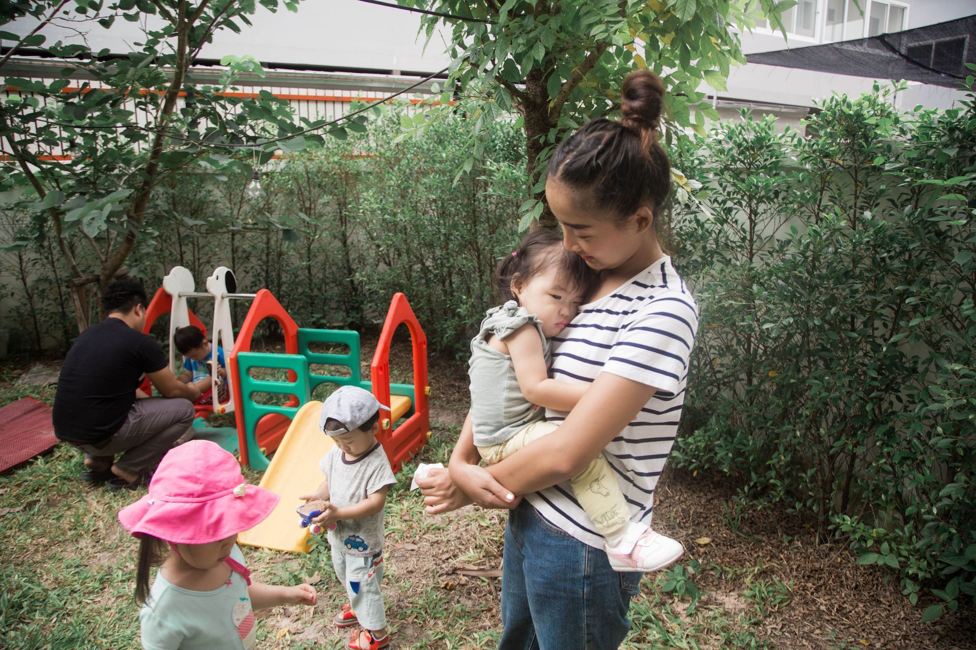 Woman holding a toddler near a playground, while two children play by a slide and hedge.