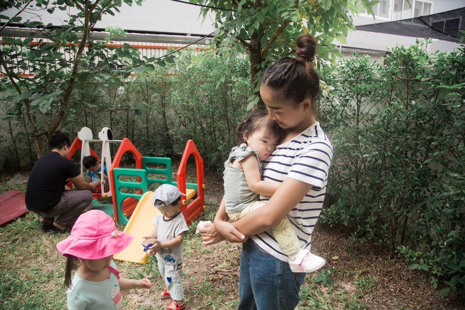Woman holding a toddler near a playground, while two children play by a slide and hedge.