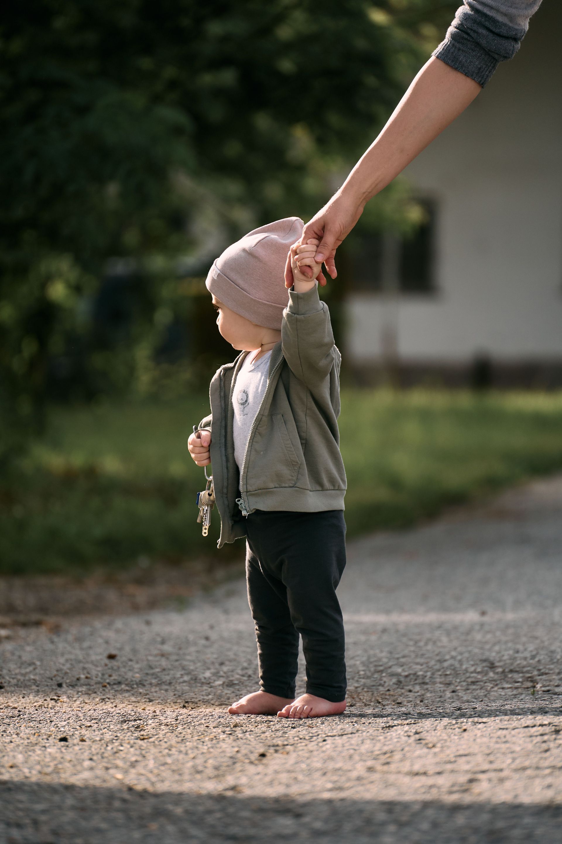 Adult holding a child’s hand outdoors on a gravel path, with a blurred building and trees in the background