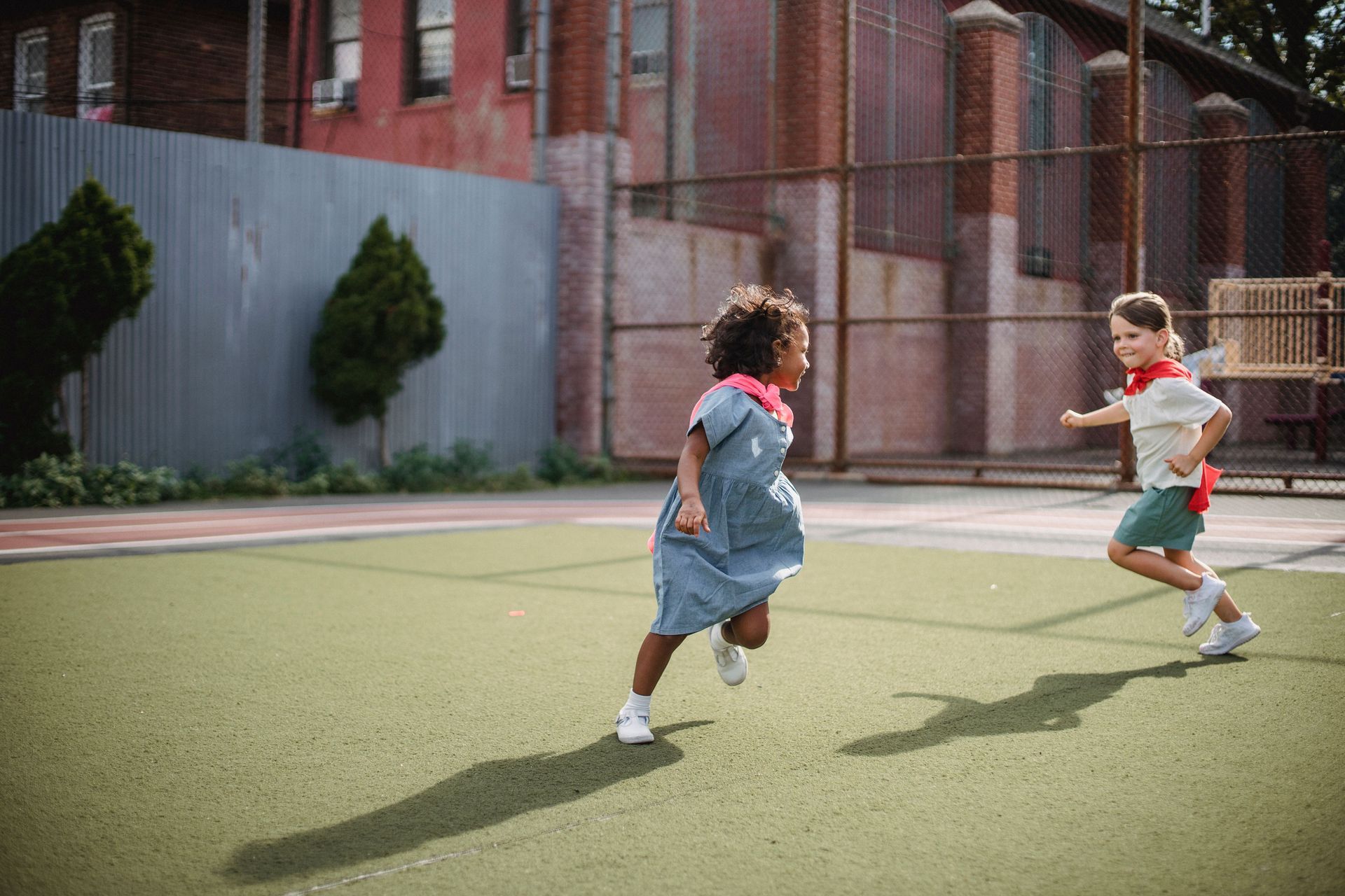 Two children running on a turf playground near a brick building and fence