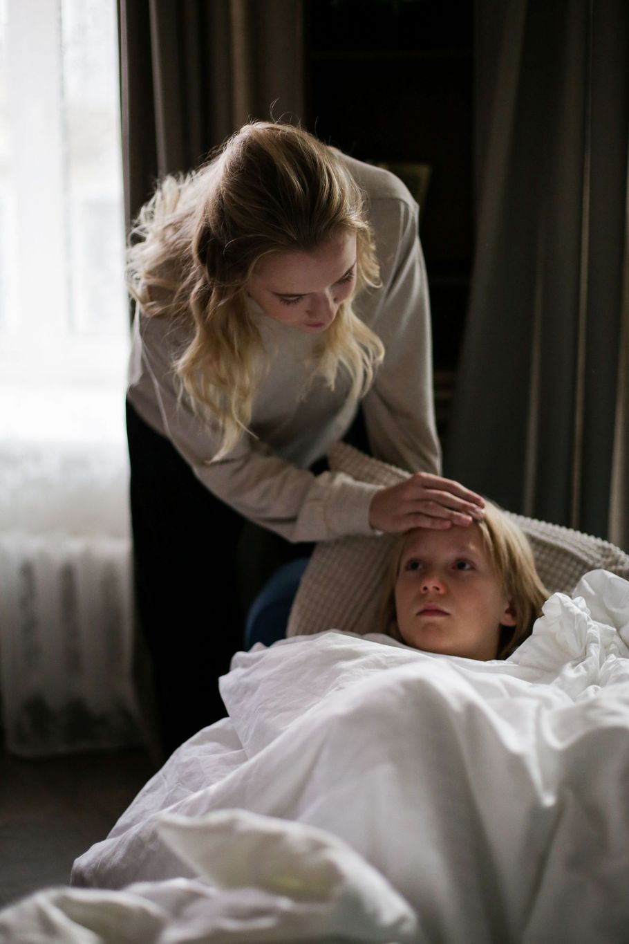 Woman gently checks another person lying in bed, in a dimly lit room.