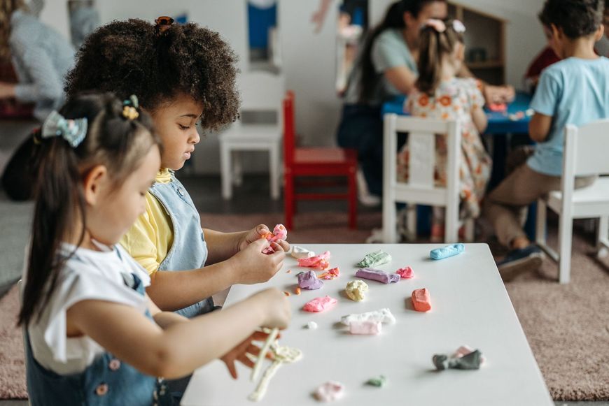 Children making crafts at a table in a classroom, with colorful paper and toy pieces scattered around.