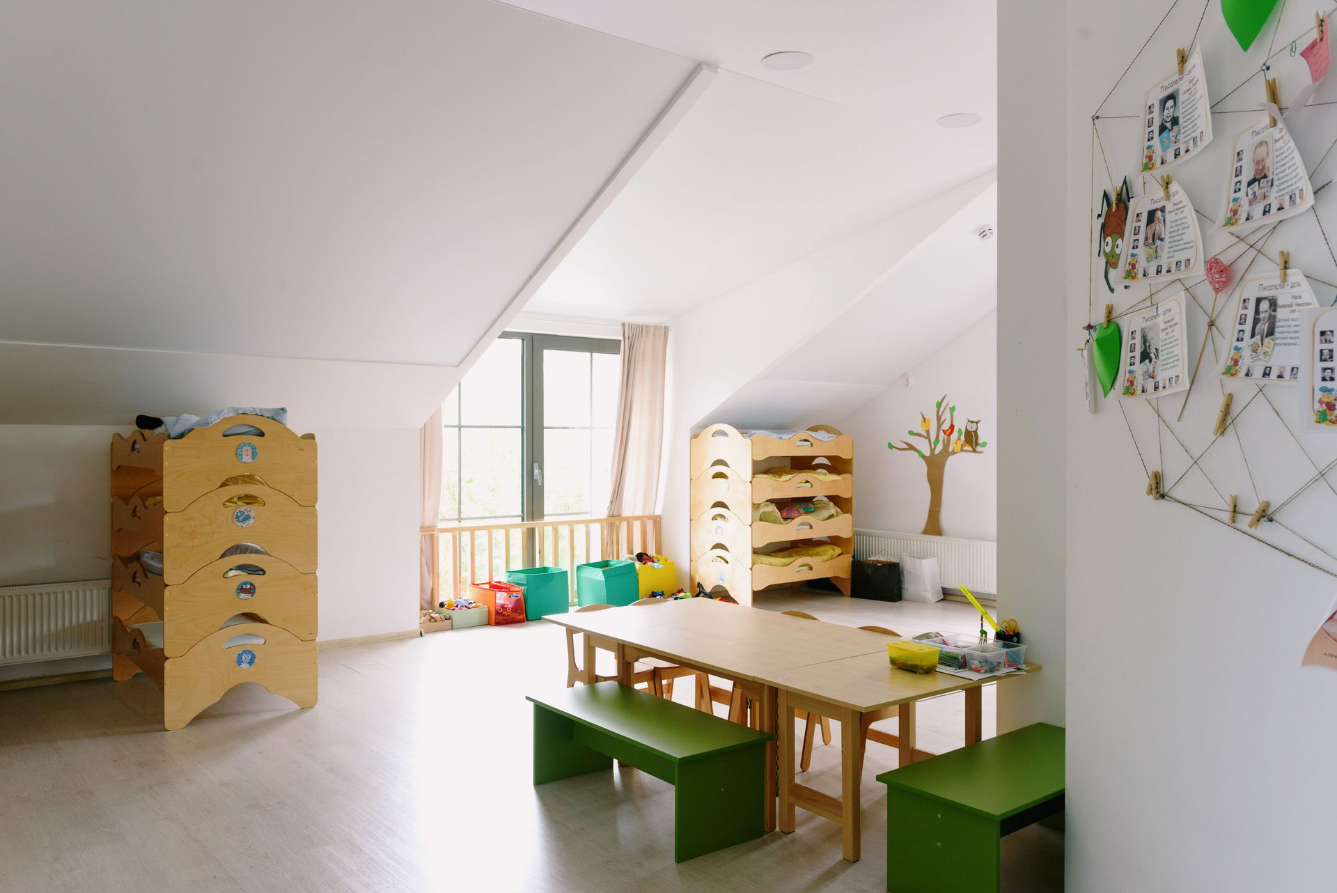Bright classroom with wooden tables, green benches, shelves, and wall art under a slanted ceiling