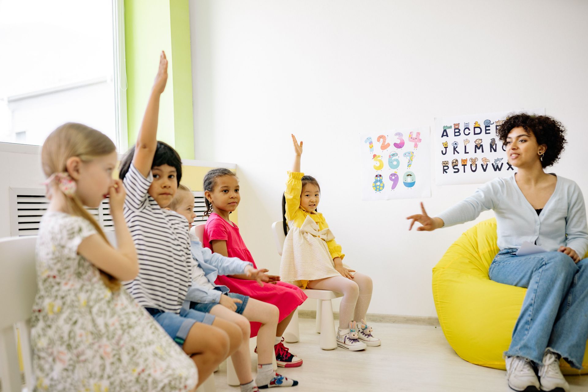 Children raising hands in a bright classroom with a teacher seated on a yellow beanbag