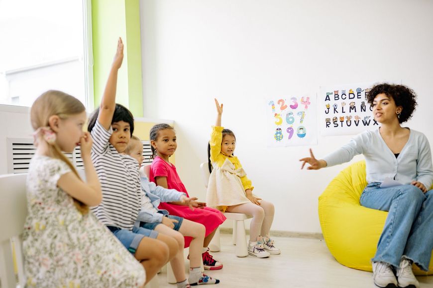 Children raising hands in a bright classroom with a teacher seated on a yellow beanbag