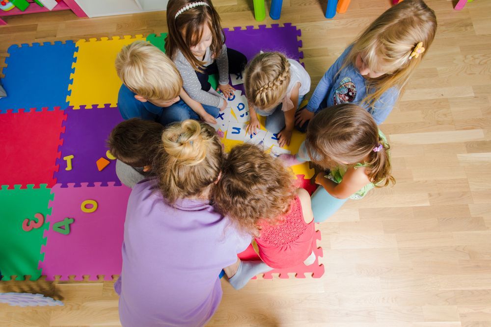 Children gathered around a table, playing with colorful toys on foam mats in a classroom.