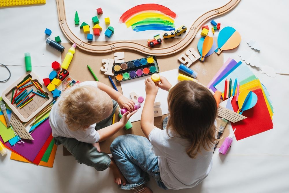 Two children painting at a table with colorful art supplies and a rainbow craft on a white floor