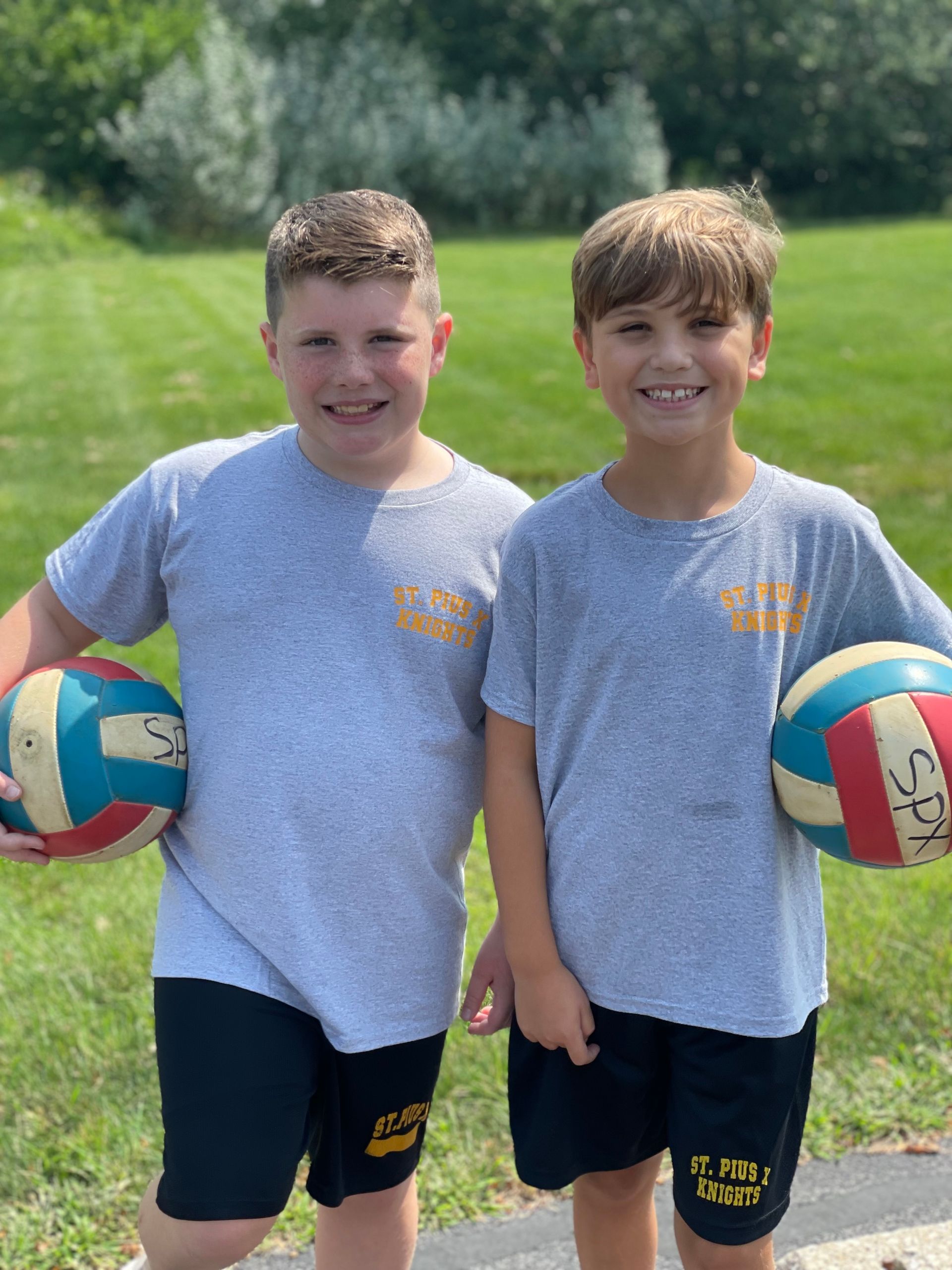 Two young boys are standing next to each other holding volleyballs.
