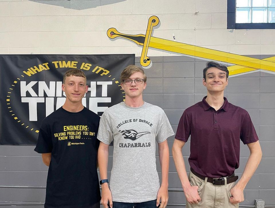 Three young men are posing for a picture in front of a knight time banner.