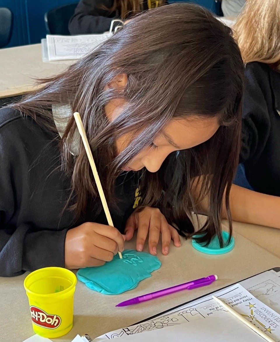 A girl is sitting at a desk with a cup of play doh and a pen.
