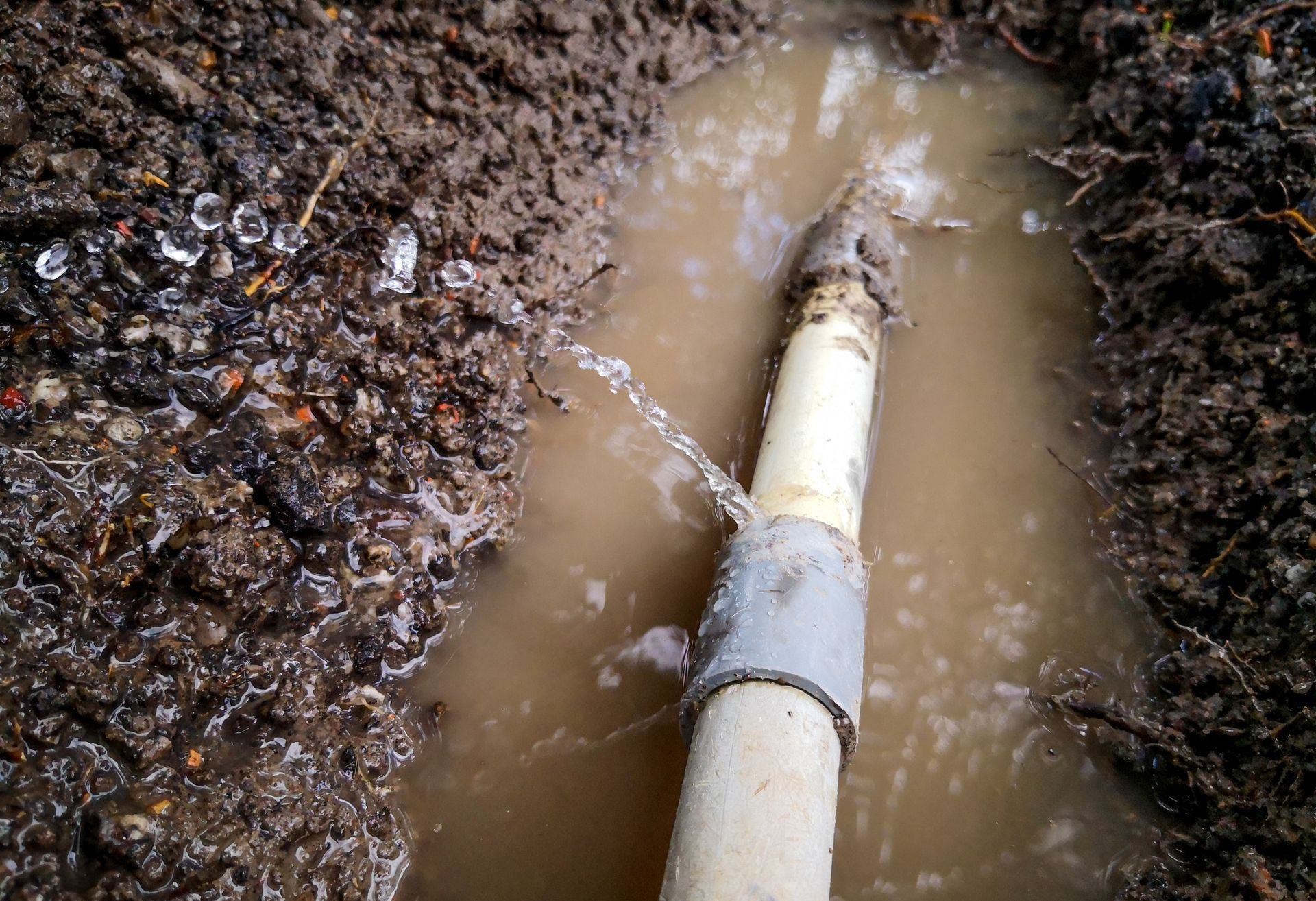 Broken white pipe leaking muddy water in a trench