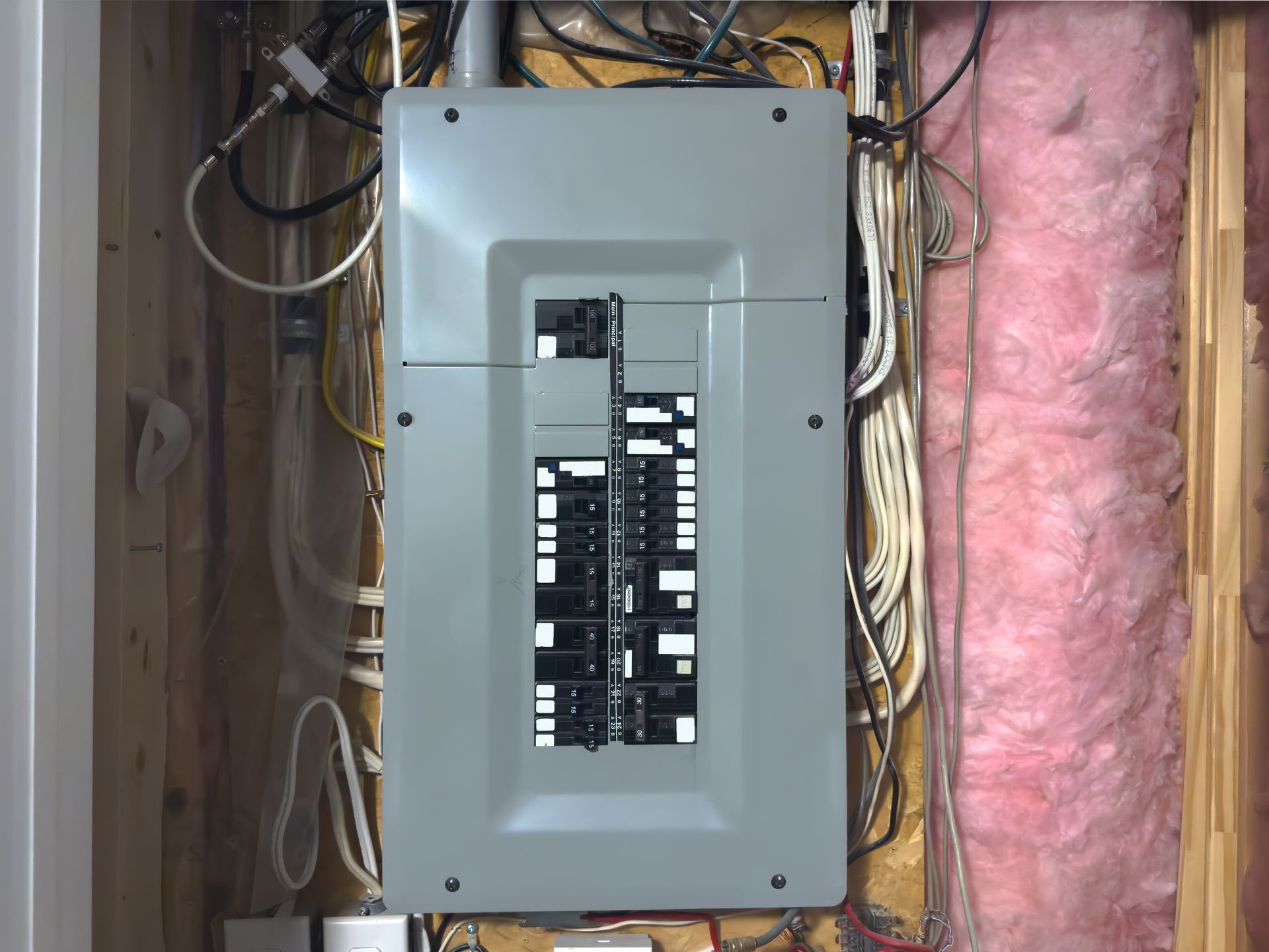 Open electrical breaker panel in a wall, surrounded by exposed wiring and pink insulation.
