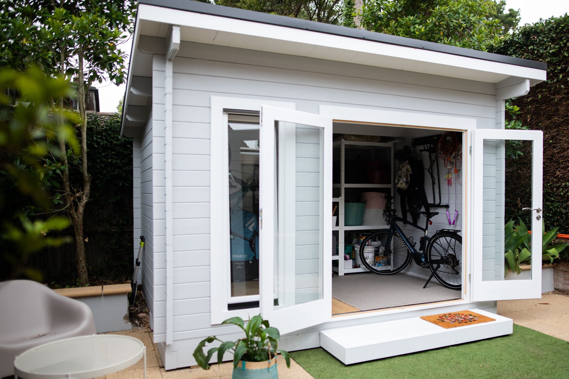 Petit hangar gris avec portes françaises blanches ouvertes, vélo à l'intérieur, sur l'herbe verte.