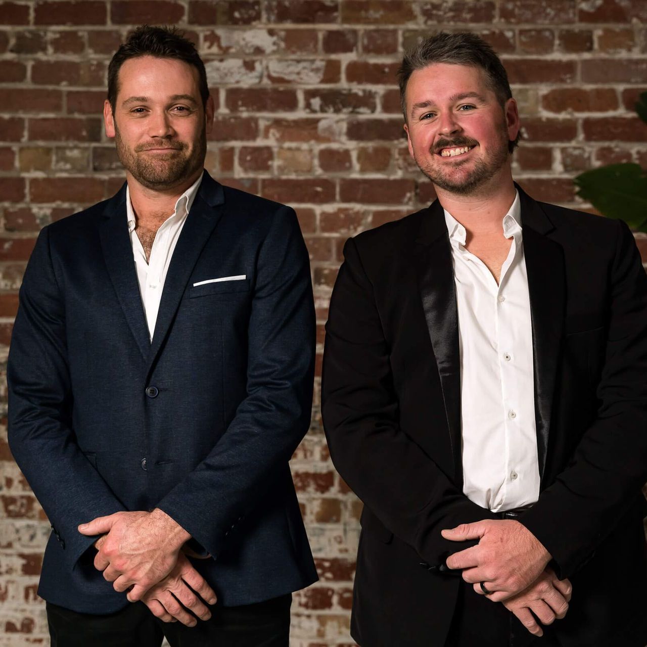 Two men in dark suits and white dress shirts stand side-by-side against a brick wall, both smiling with hands clasped.