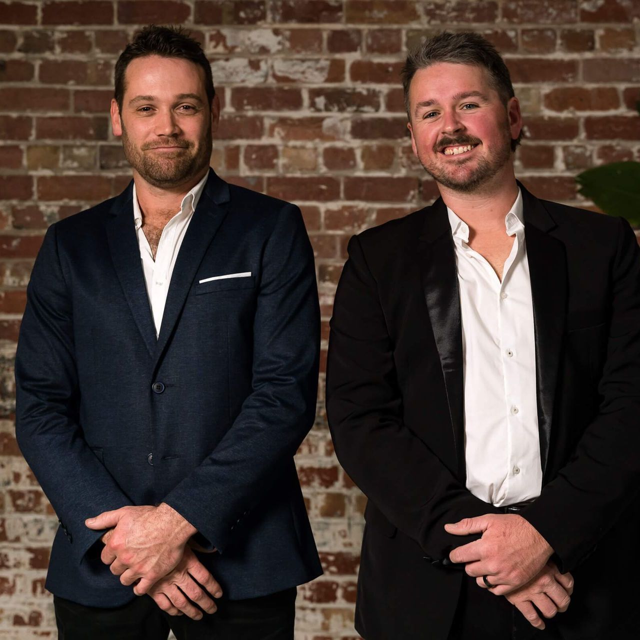 Two men in dark blazers and white dress shirts standing side by side against a brick wall, hands clasped, smiling.
