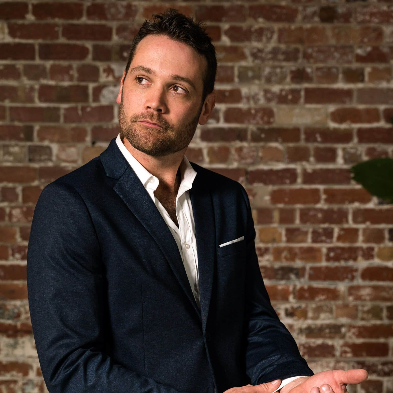 A person with a beard, wearing a white shirt and dark blue blazer, poses against a rustic red brick wall.