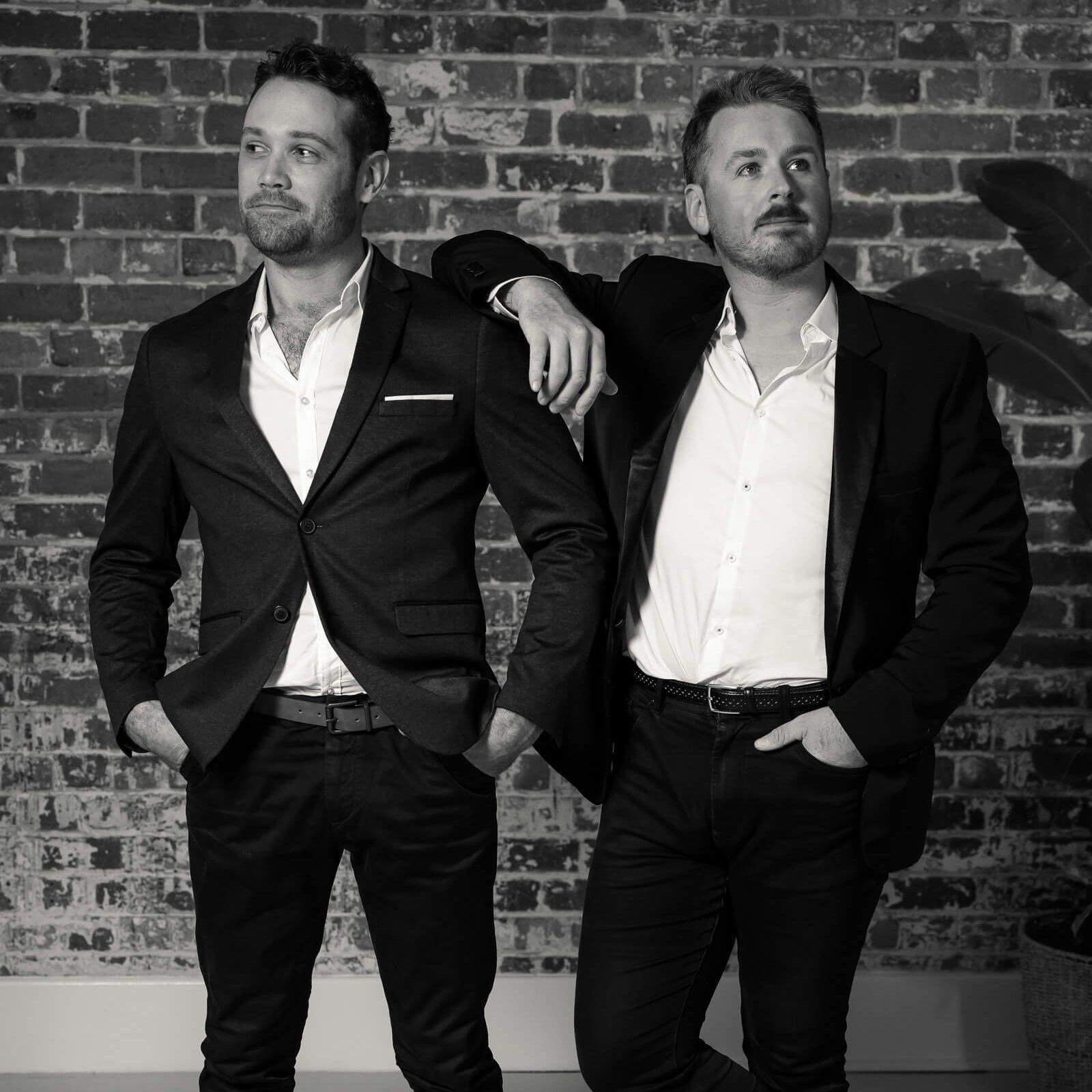 Two men in suits and open-collared shirts standing against a brick wall, one resting his arm on the other's shoulder.