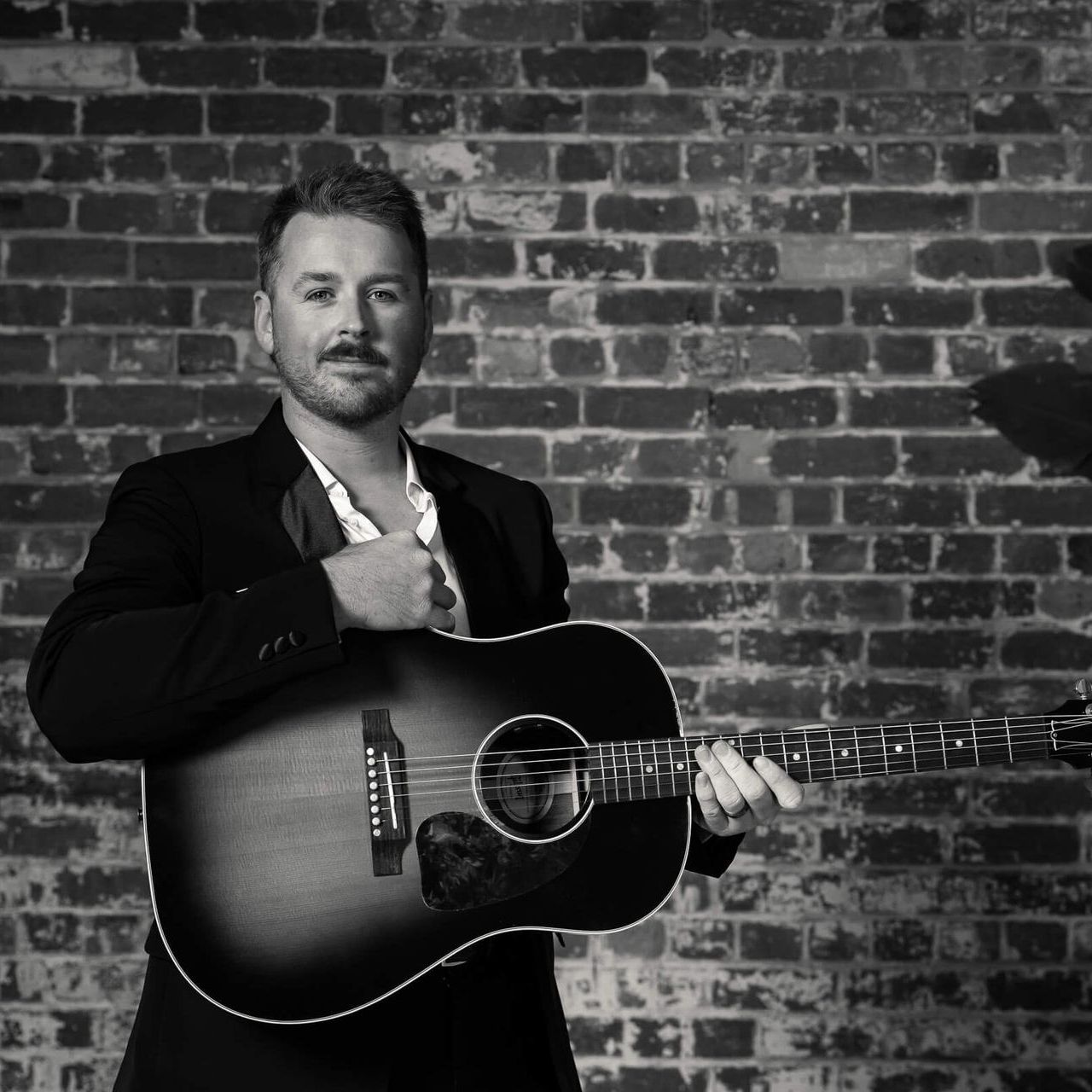 A person wearing a suit holds an acoustic guitar against a brick wall background.