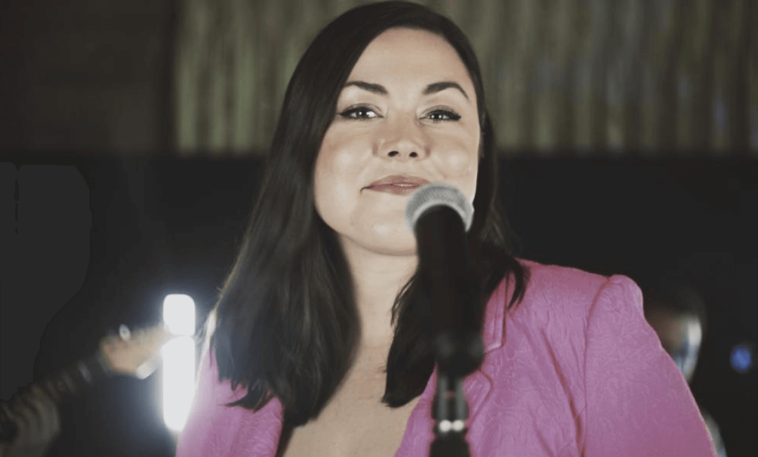 Woman singing into a microphone, wearing a pink blazer. Stage lights behind her.
