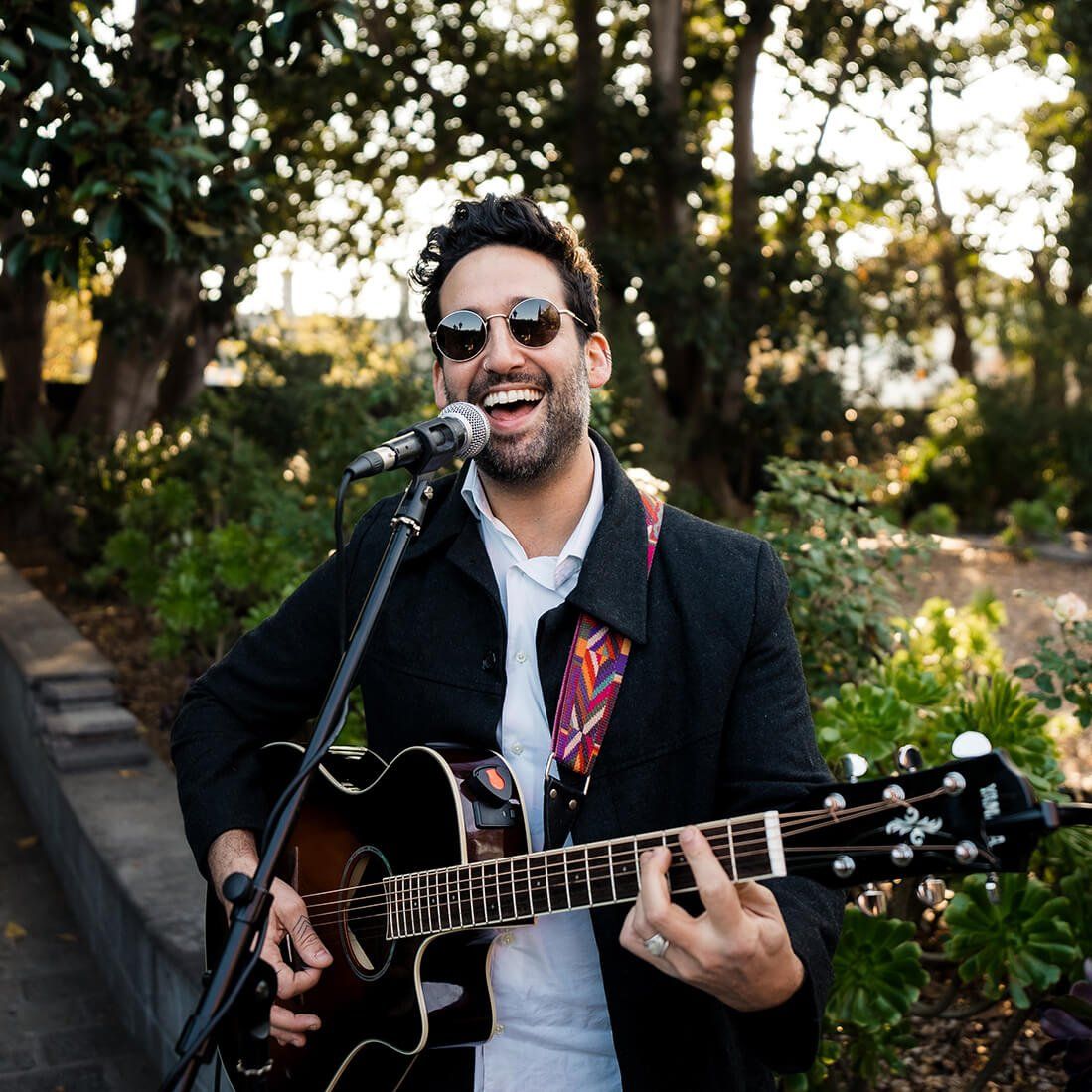 Man playing acoustic guitar and singing into a microphone outdoors; wearing sunglasses and a black jacket.