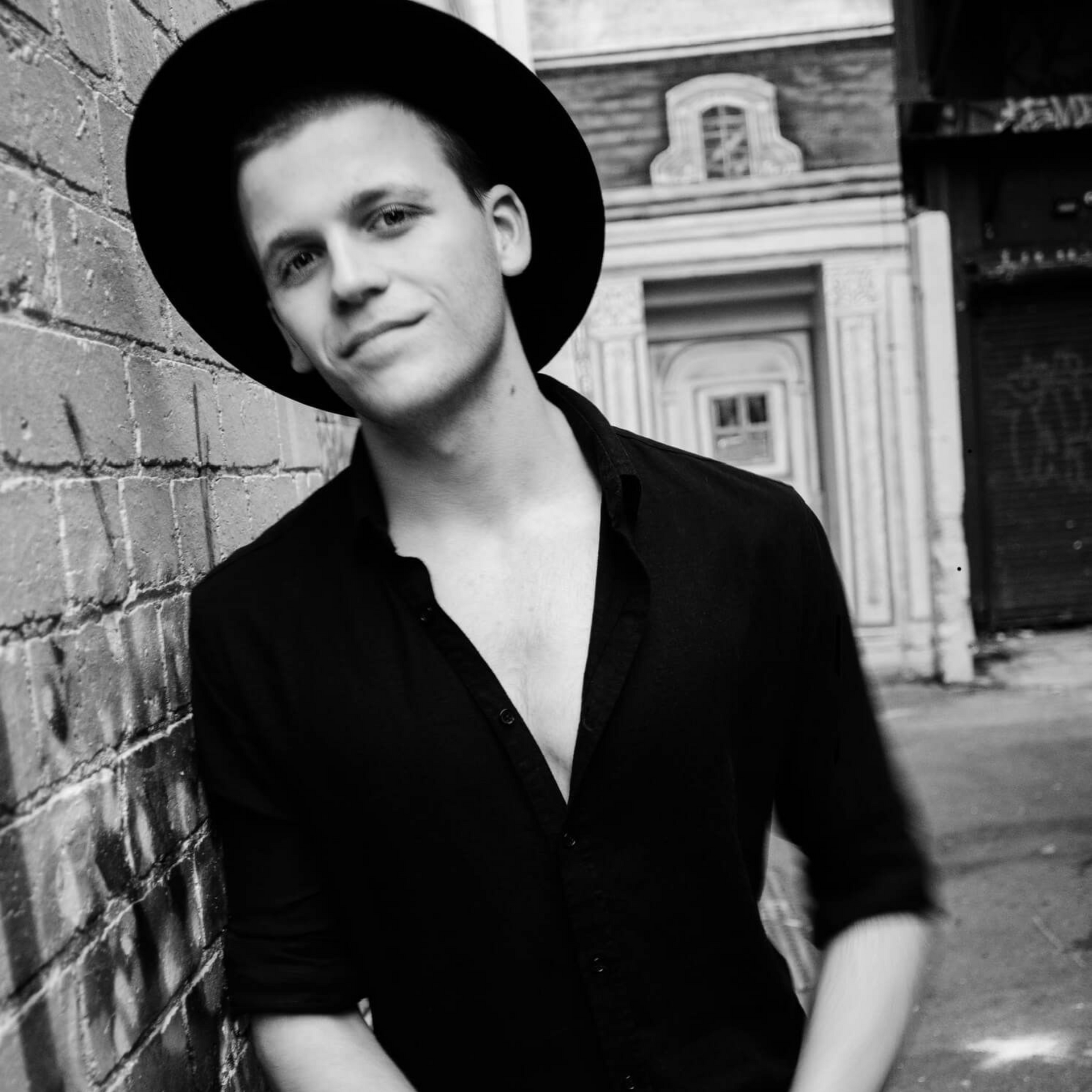 Person in a black hat and shirt leaning against a brick wall in a black-and-white street portrait