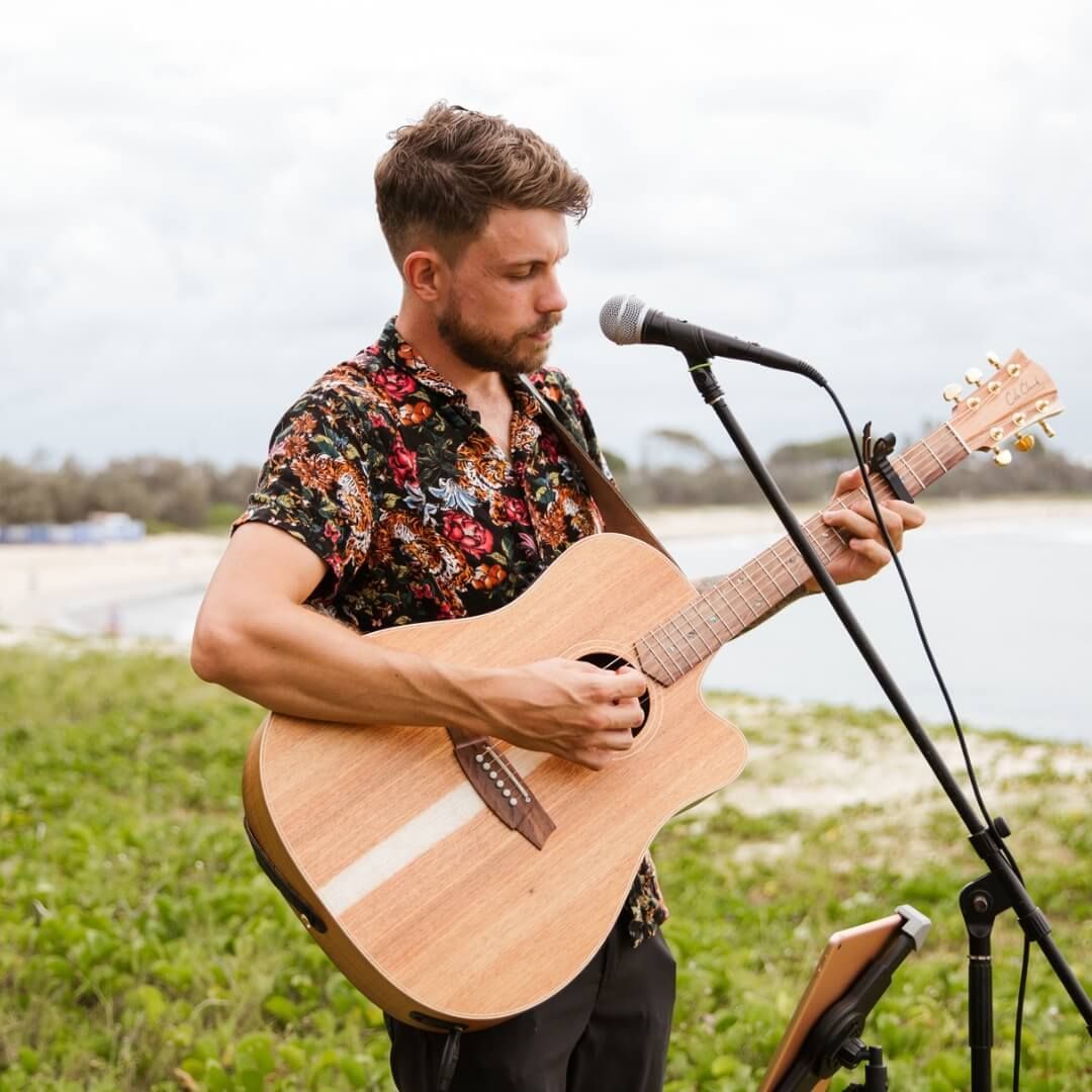 A person playing an acoustic guitar and singing into a microphone outdoors by the beach.