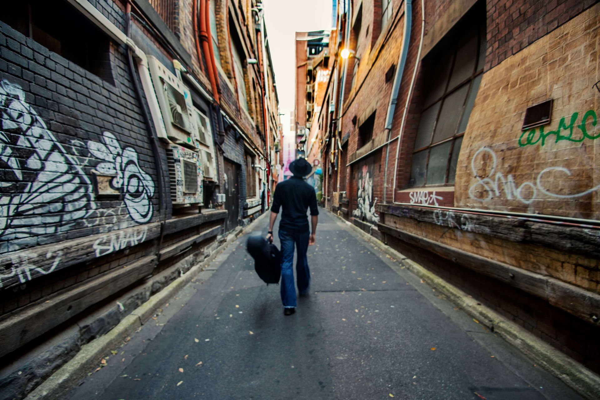 Person walking down a narrow graffiti-lined alley, carrying a black bag.