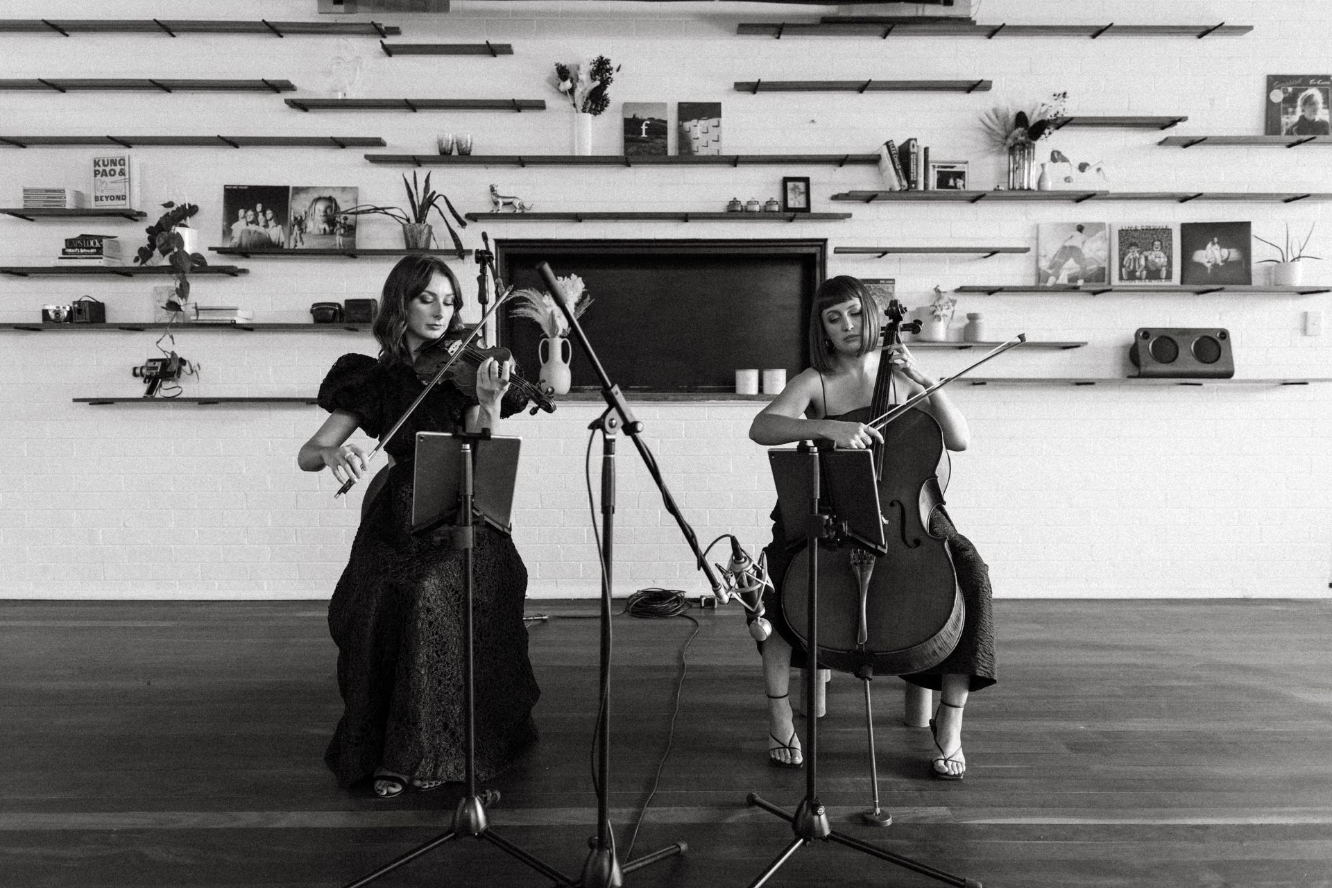 Two musicians play a violin and a cello in a room with shelving displaying various items on a white wall.
