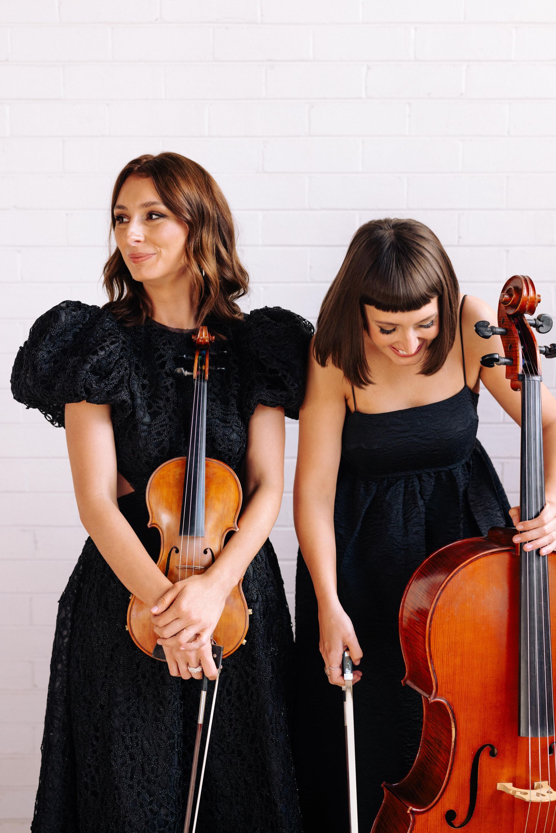 Two musicians in black dresses stand before a white brick wall, one holding a violin and the other a cello.