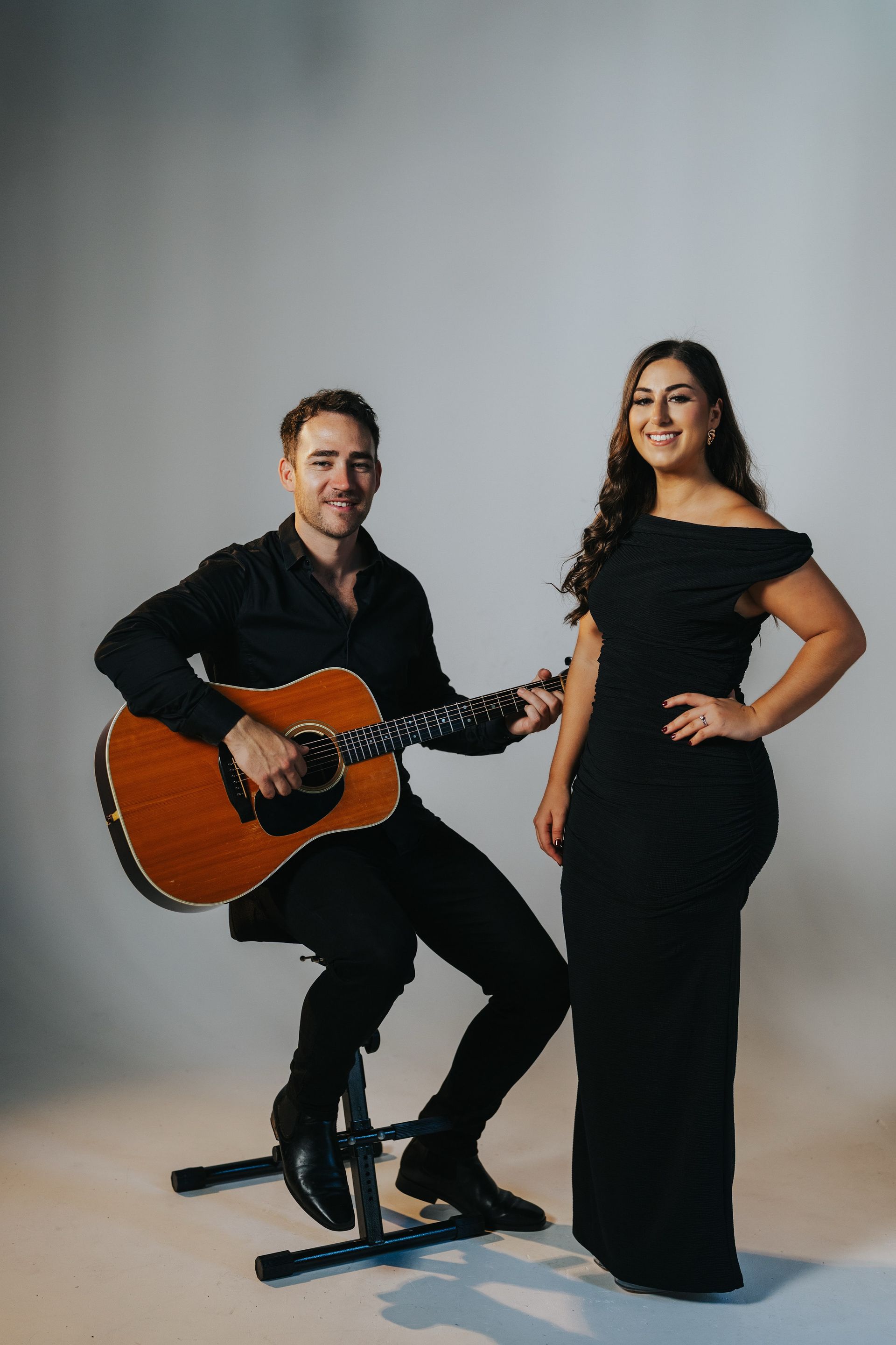 A man seated with an acoustic guitar and a woman in a black gown stand together against a light gray backdrop.