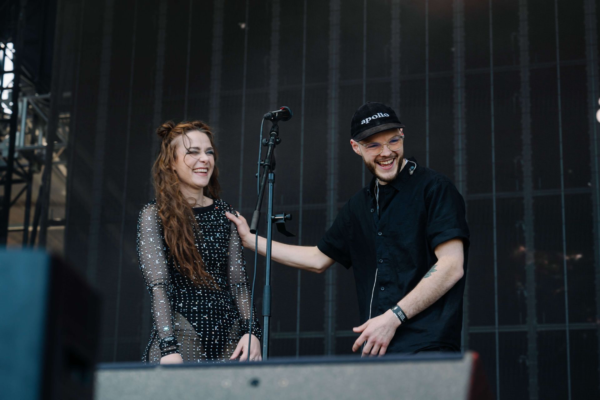 Two people smiling onstage, standing by a microphone, one adjusting it during a live performance.