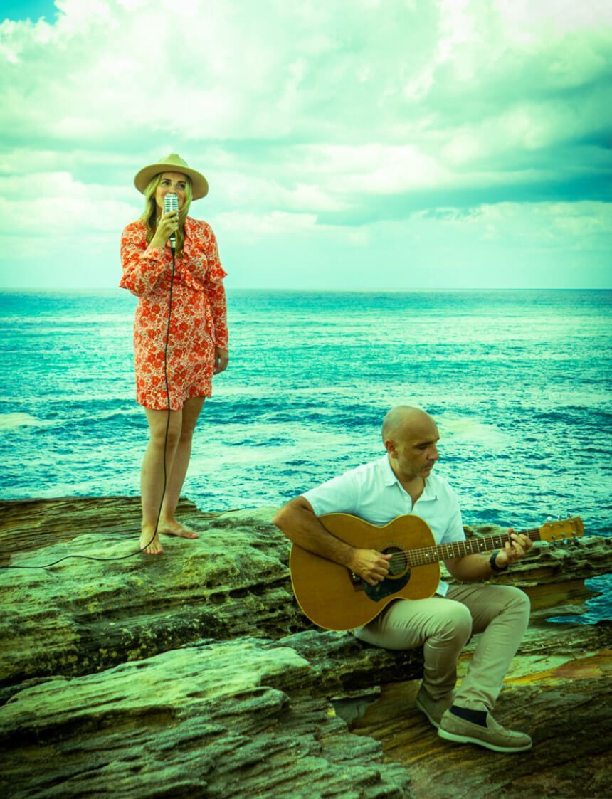 A musician plays an acoustic guitar while sitting on ocean rocks, as a singer performs into a microphone nearby.