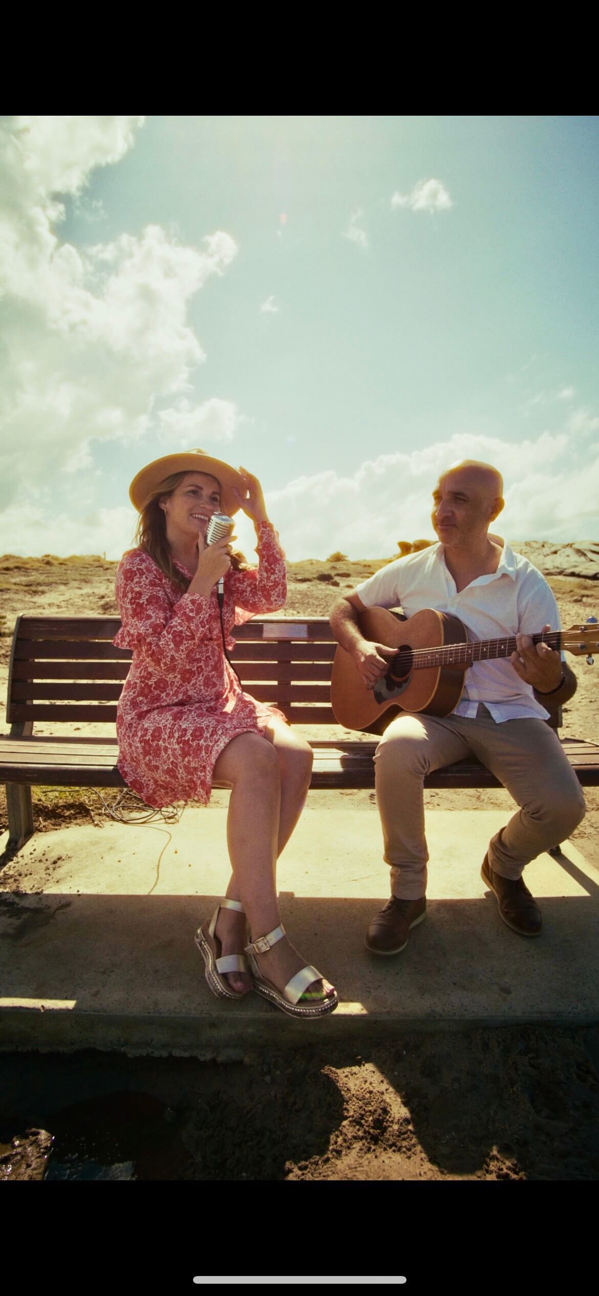 A person wearing a floral dress and straw hat sings while another person plays an acoustic guitar on a wooden park bench.