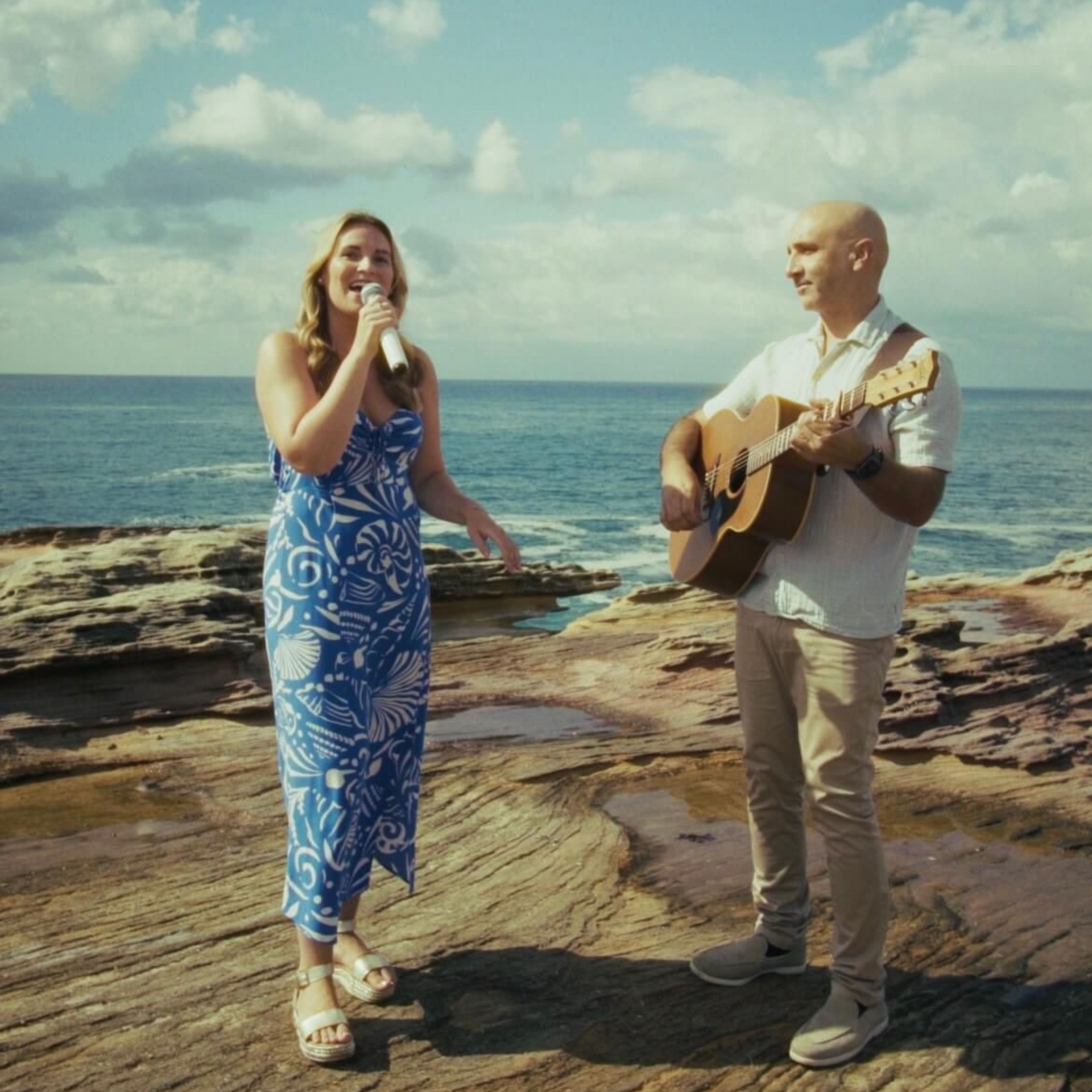 A person singing into a microphone while standing next to someone playing an acoustic guitar on a rocky beach by the sea.
