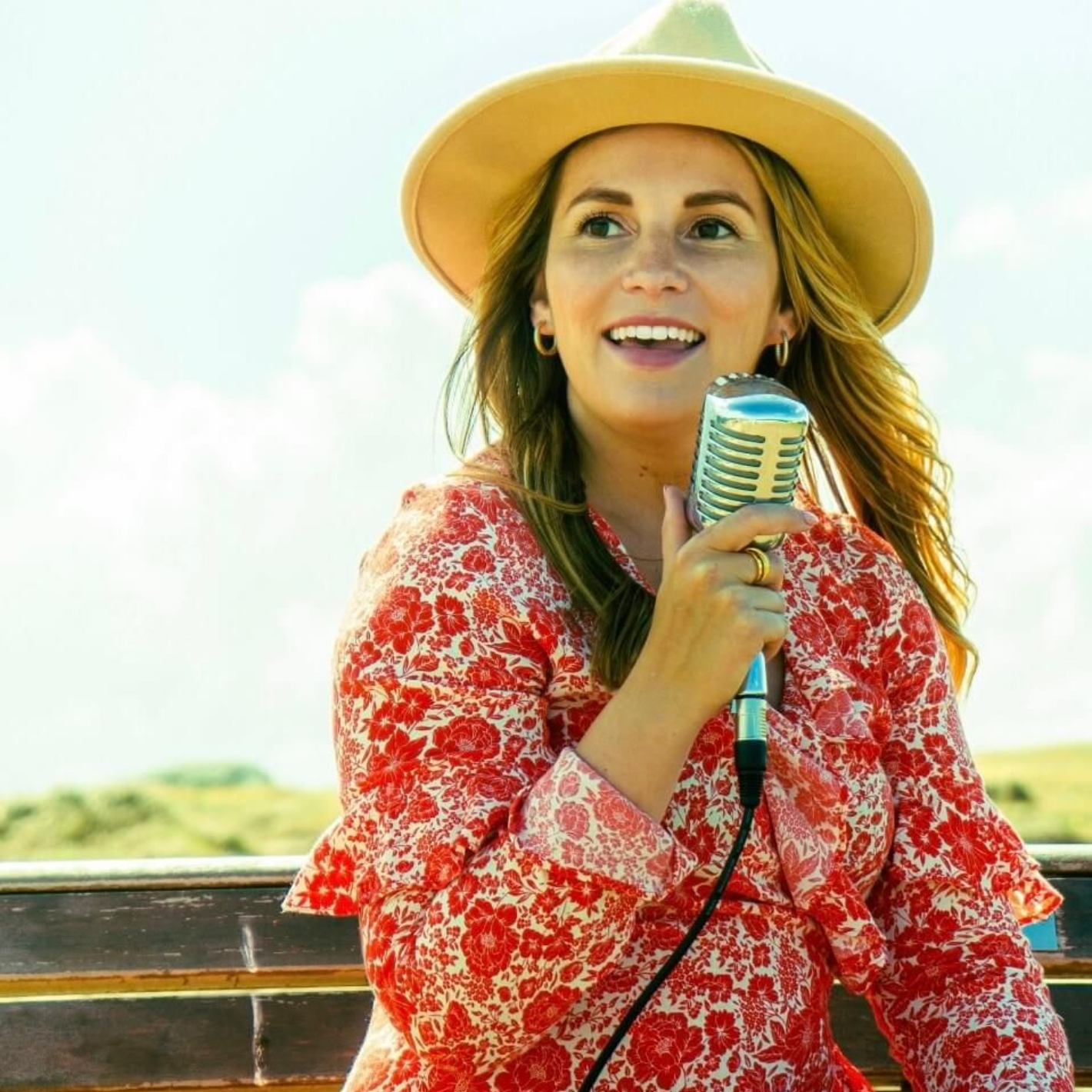 A person wearing a tan fedora and floral red dress sings into a vintage-style microphone outdoors.