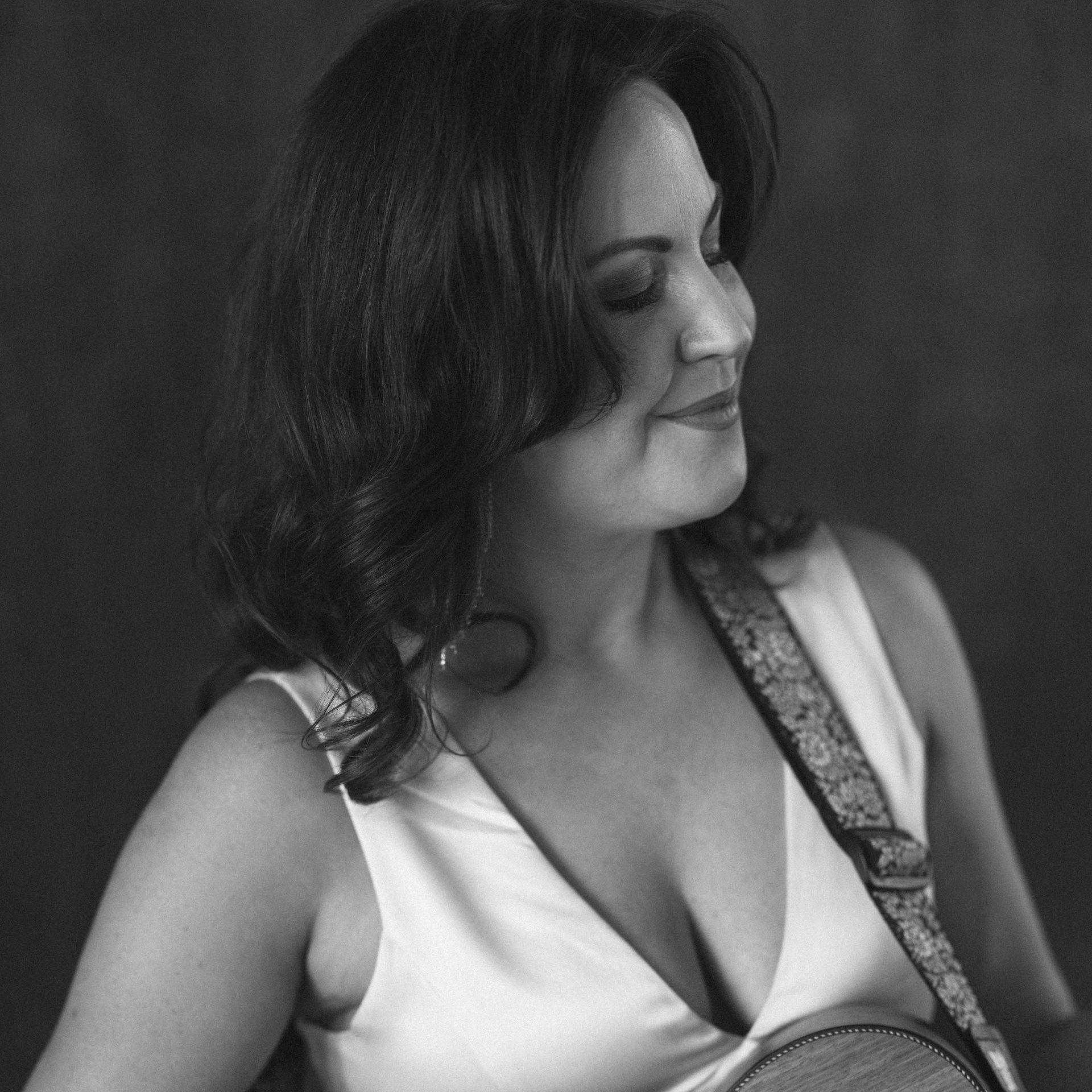 Woman in white top, holding stringed instrument, looking to the side, smiling.