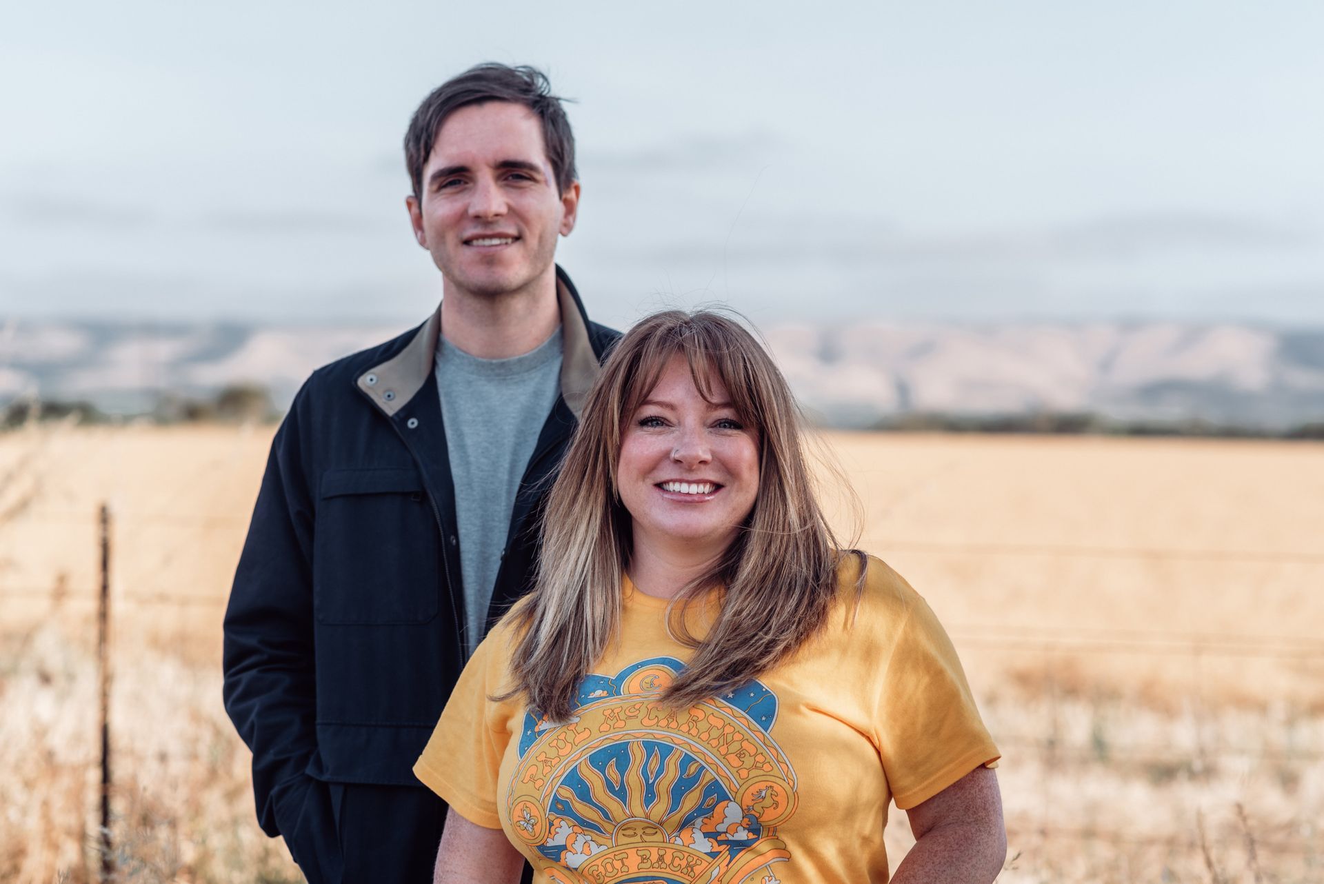 Man and woman standing outdoors, smiling. Woman in yellow shirt, man in jacket. Field and mountains in background.