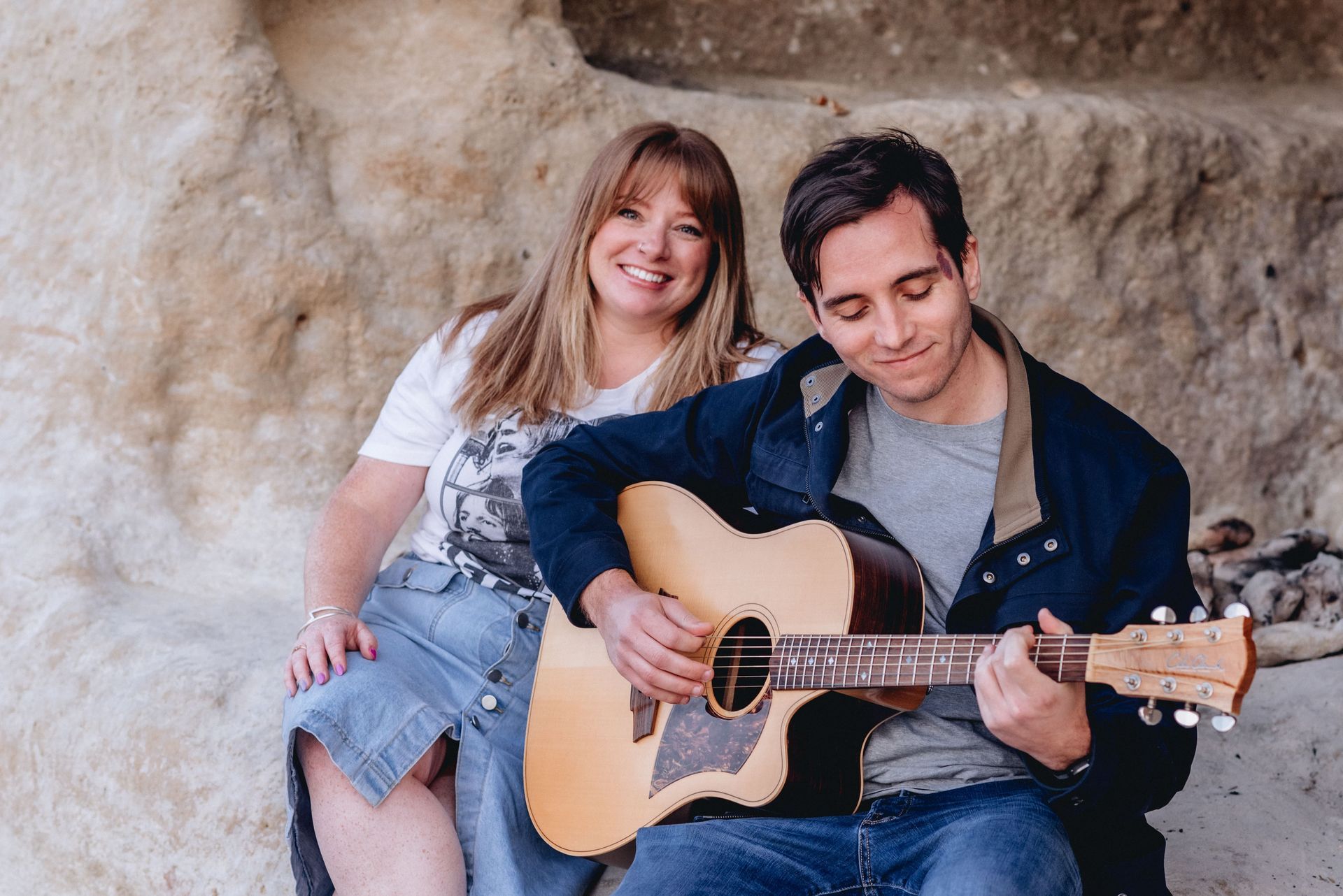 Woman and man seated near a rock wall. Man plays acoustic guitar. Woman smiles, looks toward the viewer.