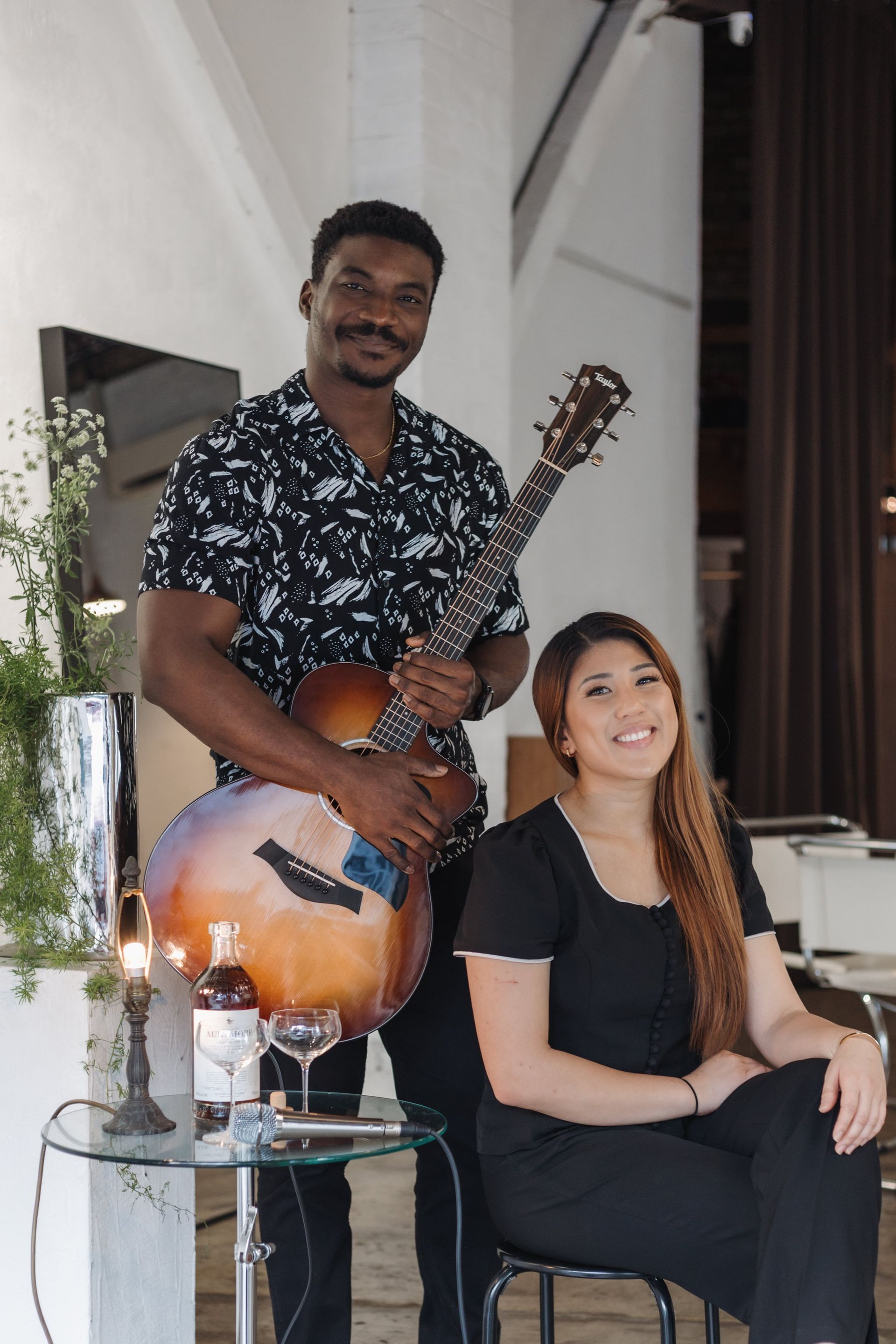 A person holding an acoustic guitar stands next to a person sitting on a stool in a bright, modern room with a drink.