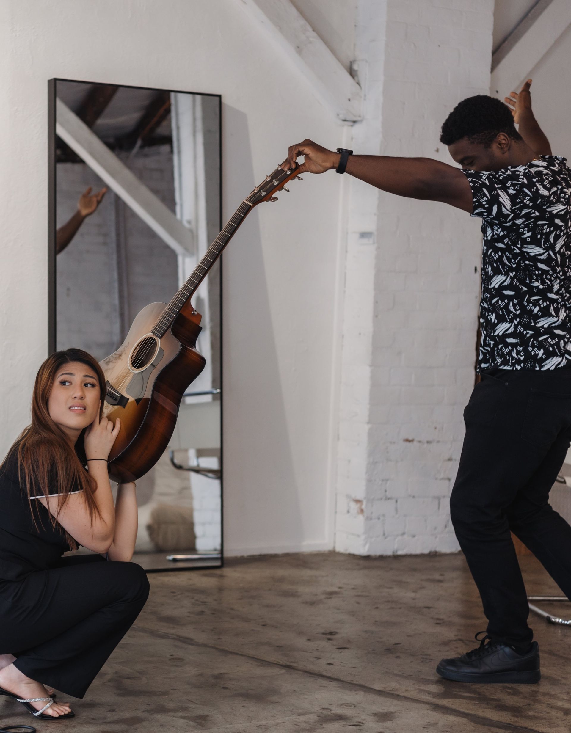 A person holding the neck of an acoustic guitar while another person crouches near a floor mirror, interacting with it.
