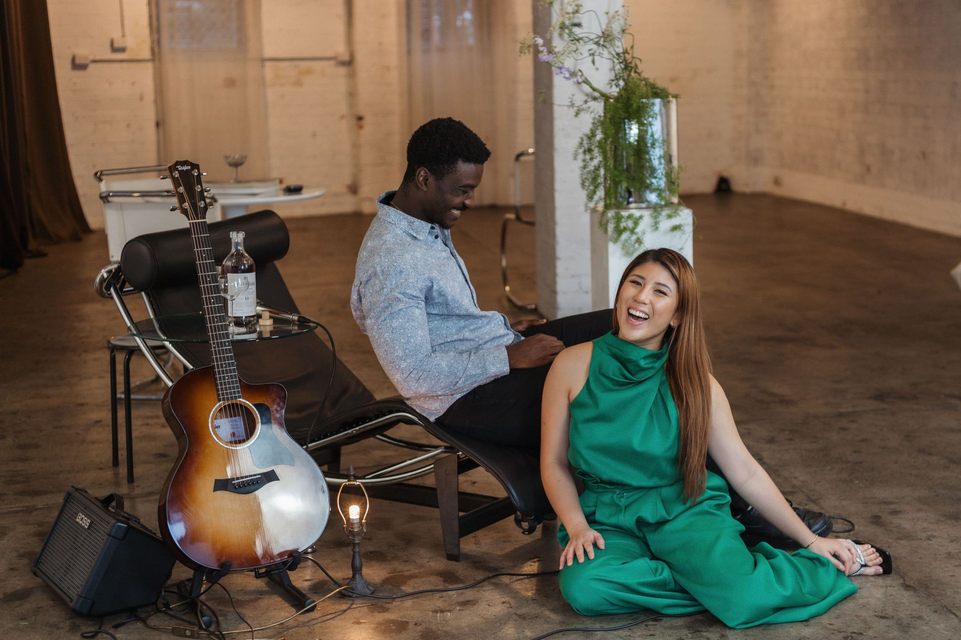 A man in a blue shirt sits on a lounge chair while a woman in a green outfit sits on the floor; an acoustic guitar is nearby.
