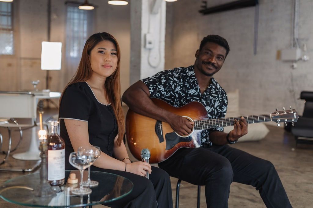 A woman and a man playing acoustic guitar in a bright, modern studio with a bottle and glasses on a glass table.