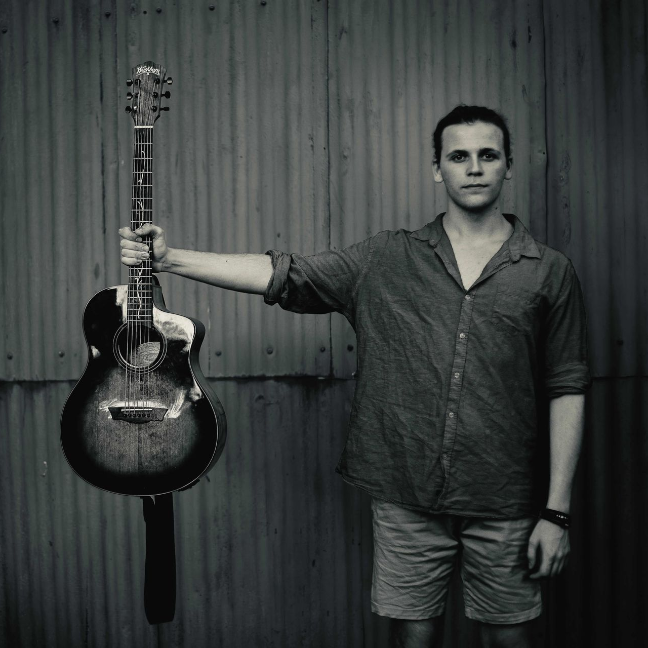 Person holding a guitar by the neck against a corrugated metal wall.