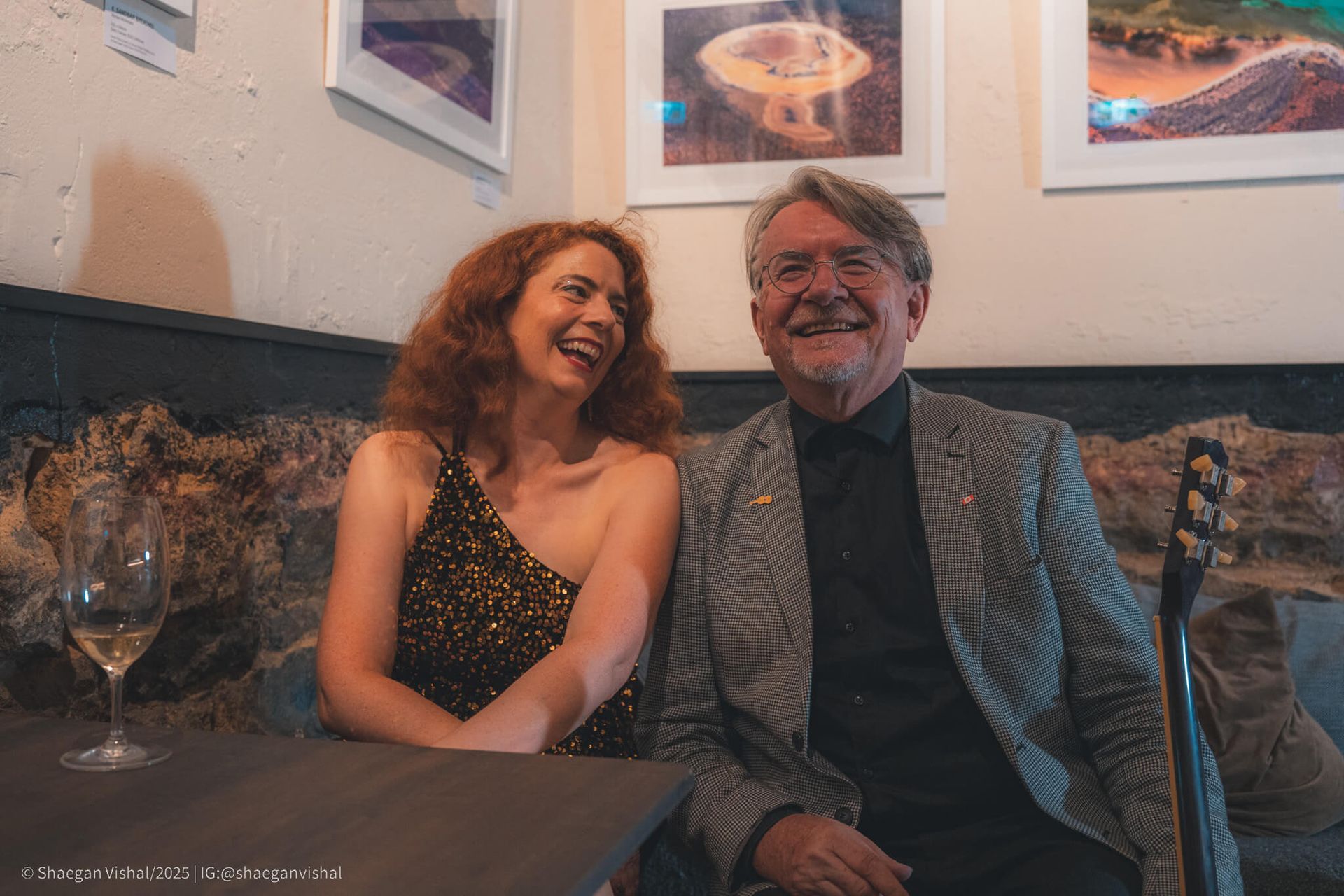 Woman with red hair and man smiling together in a dimly lit room, seated at a table.