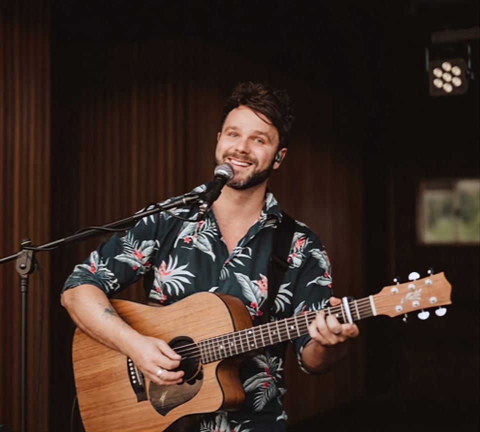 A musician with a beard smiles while playing an acoustic guitar and singing into a microphone in a dimly lit room.